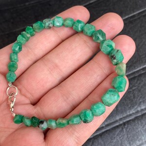 May include: A beaded bracelet featuring faceted, irregular green gemstone beads with natural dark inclusions. The beads are strung together and secured with a silver-toned metal lobster claw clasp and a small circular jump ring connector.