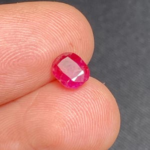 May include: A faceted, oval-shaped ruby gemstone. The ruby is a vibrant red colour and appears to be held between two fingers. The gemstone has a polished surface and reflects light, showcasing its internal structure.