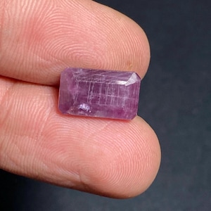 May include: A rectangular, faceted, pink gemstone. The gem is held between two fingers, showcasing its polished surfaces and internal structure. The stone's colour ranges from light to deep pink, with visible internal lines.