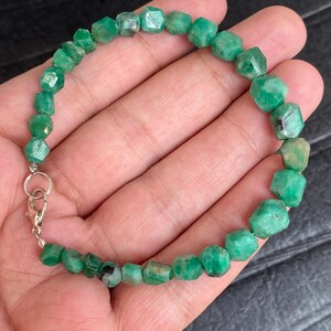 May include: A beaded bracelet featuring numerous small, faceted, vibrant green emerald stones strung together. The bracelet is secured with a silver-toned metal lobster claw clasp and a circular jump ring, creating a continuous circular loop.