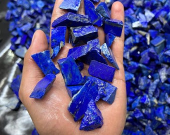 May include: Handful of raw, deep blue lapis lazuli stones. The stones are irregularly shaped, with a rough texture and varying shades of blue. The background is filled with more of the same stones.