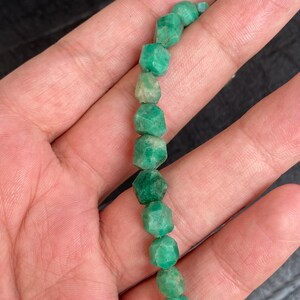 May include: A strand of small, faceted, raw emerald beads in varying shades of vibrant green. Each bead features an irregular, multi-sided shape with a matte, natural texture, strung together in a vertical line against a dark background.