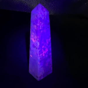 May include: A tall, four-sided obelisk-shaped crystal tower standing upright. The stone exhibits a vibrant, glowing purple and neon pink fluorescence across its entire surface, creating a mottled, textured appearance under ultraviolet light.