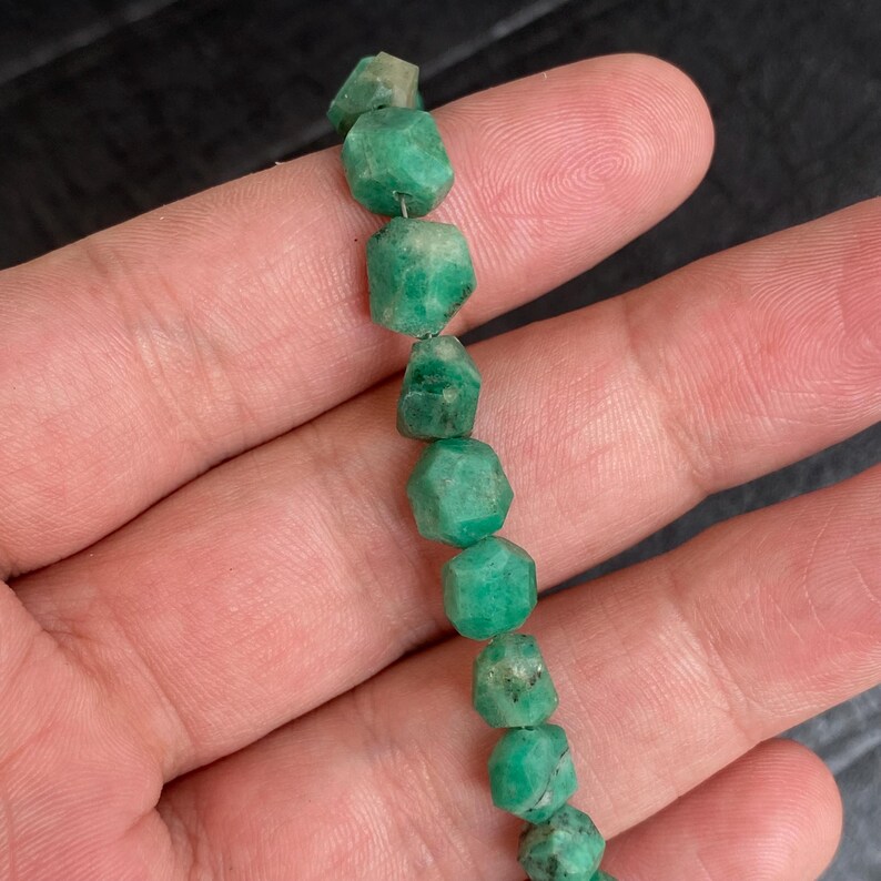 May include: A strand of raw, faceted emerald beads in varying shades of deep and light green. Each bead features an irregular, multi-sided shape with a natural, unpolished texture, strung together on a thin, light-colored cord for jewelry making.
