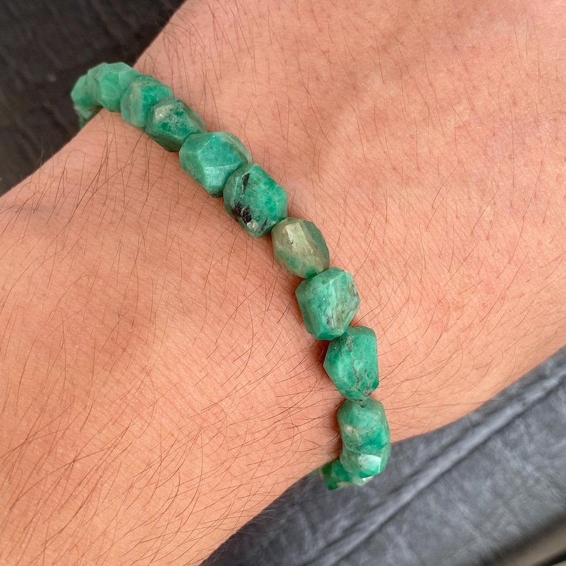 May include: A bracelet composed of faceted, irregular, raw green emerald beads strung together. The stones exhibit varying shades of deep and light green with natural inclusions and a textured, matte surface finish throughout the piece.