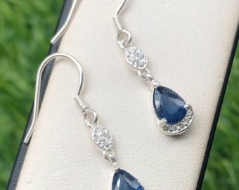 May include: A pair of silver drop earrings. Each earring features a teardrop-shaped, deep blue gemstone, surrounded by a halo of small, clear stones. Above the blue stone is a small, oval cluster of clear stones. The earrings have a hook closure.