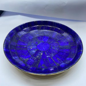 Op de afbeelding: Een cirkelvormig decoratief dienblad met een mozaïekoppervlak bestaande uit diepblauwe lapis lazuli steensegmenten. Het dienblad heeft een dunne goudkleurige metalen rand die het ingelegde steenpatroon omsluit, uitstralend vanuit het midden.