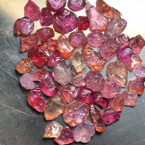 Puede incluir: Una colección de piedras preciosas en bruto y sin pulir en varios tonos de rosa, rojo, naranja y morado. Las piedras tienen formas irregulares y facetadas con un acabado brillante. El texto en la parte inferior dice: Minor Oil Added To Increase Visual Clarity.