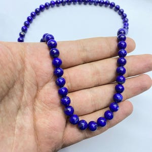 May include: A necklace composed of round, deep blue lapis lazuli beads. The beads feature subtle natural variations and dark inclusions, strung together in a continuous strand. The polished surface of each bead reflects light, highlighting the intense blue color and the spherical shape of the gemstone components.