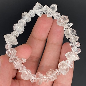 May include: A clear quartz crystal bracelet. The bracelet is composed of irregularly shaped, faceted quartz crystals strung together. The crystals are transparent and reflect light, creating a sparkling effect. The bracelet is held in a hand against a dark background.