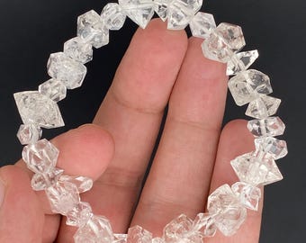 May include: A clear quartz crystal bracelet. The bracelet is composed of irregularly shaped, faceted quartz crystals strung together. The crystals are transparent and reflect light, creating a sparkling effect. The bracelet is held in a hand against a dark background.