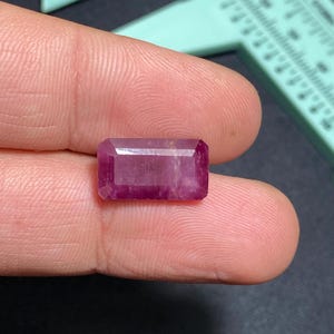 May include: A rectangular, faceted ruby gemstone with a deep pinkish-red hue. The gem is held between fingers, showcasing its clarity and cut. The background includes a measuring tool, suggesting a focus on precision and detail in the gem's presentation.