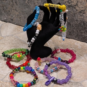 May include: A collection of colorful beaded bracelets with various words and phrases. The bracelets are displayed on a black hand-shaped stand. The bracelets feature words like "ORAL", "PAY", "FUCK", and "WHERE'S MOLLY".