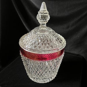 May include: A clear glass candy dish with a diamond pattern and a ruby red band around the middle. The lid has a pointed finial. The dish is on a black surface.