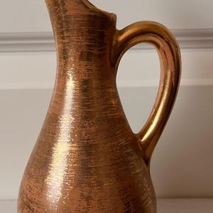 May include: A copper-colored pitcher with a slender, elongated spout and a rounded body. The pitcher has a textured surface with gold accents and a curved handle. The design is reminiscent of vintage or antique decor.