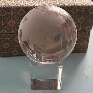 Puede incluir: Un globo terráqueo de cristal transparente con continentes grabados descansa sobre un soporte rectangular transparente. El globo está frente a una caja decorativa con un diseño marrón y dorado. Es un artículo decorativo.