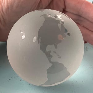 Puede incluir: Un globo terráqueo de vidrio esmerilado con una representación detallada de los continentes. El globo es de color gris claro y parece estar hecho de un material translúcido. Los continentes son ligeramente más oscuros, creando un contraste sutil. Tema: geografía y decoración del hogar.