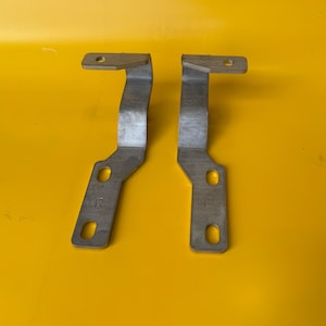 Puede incluir: Dos soportes metálicos plateados con un diseño doblado y angular. Cada soporte tiene una parte superior rectangular con un agujero y un cuerpo curvado con dos agujeros. La letra "R" está estampada en un soporte y "L" en el otro.