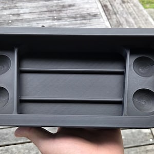Könnte beinhalten: Ein dunkelgrauer, rechteckiger Organizer mit zwei Getränkehaltern an jedem Ende und zwei zentralen Fächern. Der Organizer wird von einer Hand gehalten und scheint aus einem strapazierfähigen Material zu bestehen.
