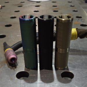 Può includere: Tre oggetti cilindrici in metallo con superfici strutturate. Uno è verde iridescente, uno nero e il terzo bronzo. Ognuno ha un'apertura circolare nella parte superiore e una fascia al centro. L'attrezzatura per la saldatura è visibile sullo sfondo.