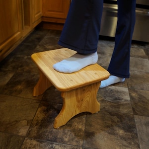 May include: A small, wooden step stool with a square top and curved legs. The stool is made of light-colored wood and appears to be in a kitchen setting. A person's foot is resting on the stool.