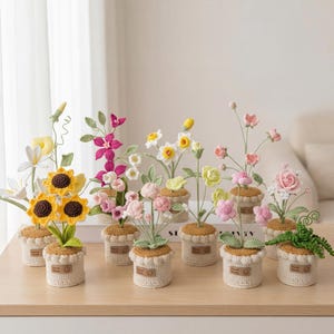 May include: A selection of handmade crochet flowers in small pots. The flowers include sunflowers, roses, and other colourful blooms. Each pot is a light beige colour with a brown trim and a small tag that says "Hand Made".