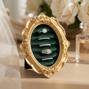 May include: A gold-colored, ornate oval ring holder with a dark green velvet interior. The holder displays five rings of varying designs, including a solitaire and several bands. The frame is detailed with floral and scroll patterns.