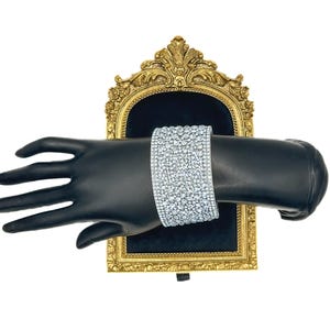 May include: A wide cuff bracelet featuring multiple rows of clear, sparkling rhinestones set in a silver-toned metal base. The bracelet is displayed on a black mannequin hand, which is positioned in front of an ornate, gold-colored frame.