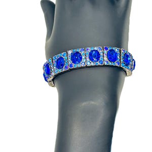 May include: A silver-toned bracelet with large, square-set, royal blue gemstones. The bracelet is adorned with smaller, iridescent blue and purple stones around the larger gems. The bracelet is displayed on a black mannequin hand against a white background.