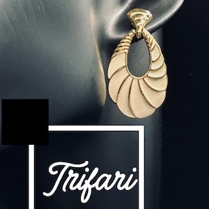 May include: Gold-toned Trifari earring with a teardrop design. The earring features a textured top and a cream-coloured, fan-like pattern within the teardrop shape. The Trifari logo is visible in the lower left corner.