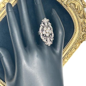 Può includere: Un anello in metallo color argento caratterizzato da un design in filigrana allungato e ornato, decorato con molteplici strass trasparenti e scintillanti disposti in un motivo floreale. L'anello presenta una finitura lucida.