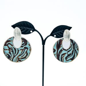 May include: A pair of turquoise and brown patterned earrings with silver-colored accents. The earrings are circular with a unique design and are displayed on a black earring stand against a white background.