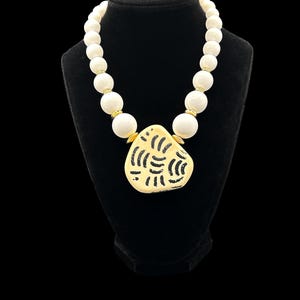 May include: A necklace featuring a strand of smooth, round, off-white beads interspersed with small, polished gold-tone spacers. The centerpiece is a large, irregular, cream-colored pendant decorated with a black, textured, curved line pattern.