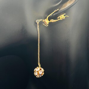 May include: A gold-colored earring with a dangling chain and a round, crystal-studded ball. The earring is designed to be worn on the ear, with a decorative element that appears to hook over the ear.
