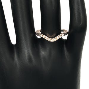 May include: A V-shaped ring featuring a metallic band with a series of small, clear, round-cut stones set along the front curve. The design follows a chevron silhouette, providing a pointed center detail that sits flush against the finger.