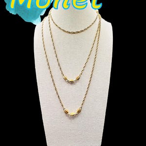 May include: A three-strand gold-tone necklace set on a white display. The necklaces have delicate chains with oval-shaped accents. The word "Monet" is written in yellow letters with a blue background.