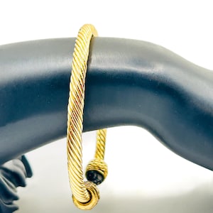 May include: A gold-colored bangle bracelet with a twisted rope design. The bracelet features a black accent at the clasp. The jewelry is displayed on a black mannequin arm against a white background.