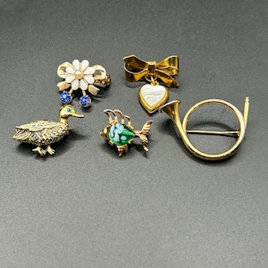 Może przedstawiać: Kolekcja broszek w stylu vintage o różnych kształtach i wzorach. Obejmuje złotą kaczkę, pierścionek z kwiatem z niebieskimi akcentami, kokardę z wisiorkiem w kształcie serca, rybę i róg. Broszki są umieszczone na ciemnym tle.