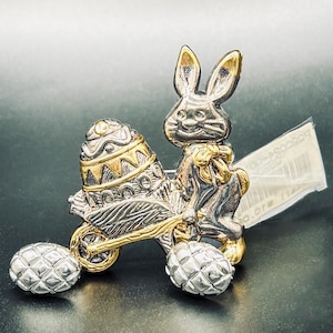 May include: A silver and gold-toned Easter brooch featuring a bunny pushing a cart with an egg-shaped wheel. The egg-shaped wheel is decorated with gold accents. Two silver egg-shaped wheels are on the ground.