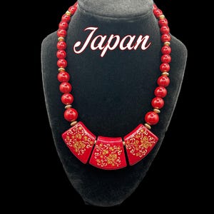 Peut inclure: Un collier rouge à perles avec trois pendentifs carrés rouges. Chaque pendentif présente un motif floral doré. Le collier est exposé sur un mannequin noir et le mot "Japan" est écrit en cursive blanche.