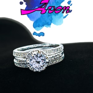 May include: A silver-colored ring set with a large, clear, round center stone and smaller stones around the band. The word "Avon" is written in pink letters above the ring, set against a blue and purple watercolor background.