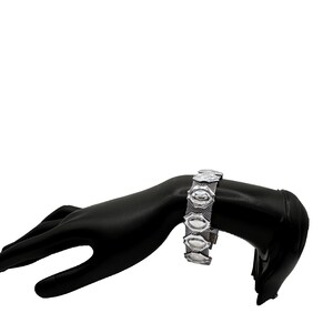 May include: Silver-toned bracelet featuring a mesh band and oval-shaped decorative elements. The bracelet is displayed on a black mannequin hand against a white background. The bracelet's design is symmetrical and the metal has a polished finish.