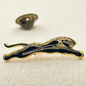 May include: A gold-toned metal lapel pin shaped like a leaping panther or cheetah. The design features a sleek black enamel fill across the body with gold-colored outlines and textured spots along the back. A separate circular metal pin back is visible.