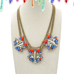 May include: A double-strand brass chain necklace featuring three ornate clusters of multi-colored rhinestones. The clusters contain bright orange, deep blue, pale green, and clear rectangular and marquise-cut crystals arranged in a symmetrical pattern.
