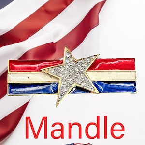 May include: A patriotic brooch featuring a star-shaped design encrusted with rhinestones, set atop a bar with red, white, and blue enamel stripes. The word "Mandle" is displayed in red text below the brooch, with a small American flag graphic.