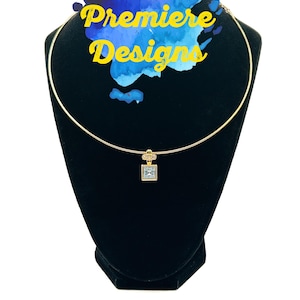 May include: A gold-colored necklace with a square, light blue pendant is displayed on a black mannequin. The necklace has a delicate chain. The text "Premiere Designs" is visible in yellow with a blue watercolor background.