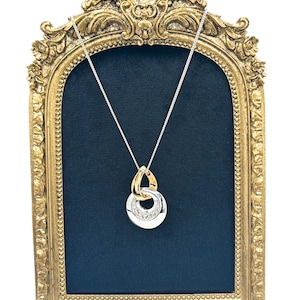 May include: A silver necklace with a gold pendant featuring a circular design with embedded crystals. The pendant is connected to a delicate silver chain. The necklace is displayed against a dark blue background within a gold ornate frame.
