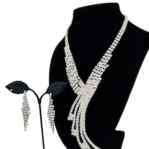 May include: A silver-toned necklace and matching drop earring set featuring clear, faceted rhinestones arranged in a multi-strand, cascading design. The necklace displays a sweeping, asymmetrical pattern, while the earrings mirror the style.