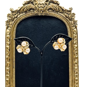 May include: A pair of gold-toned earrings with three faux pearls arranged in a cluster design. The earrings are displayed on a black leaf-shaped stand against a dark background, framed by a gold ornate decorative frame.