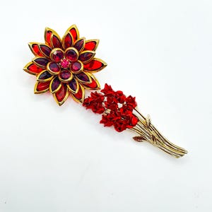 May include: Two vintage brooches. One is a large, gold-toned flower with red and purple crystals. The other is a gold-toned bouquet of red flowers. Both are set against a white background.